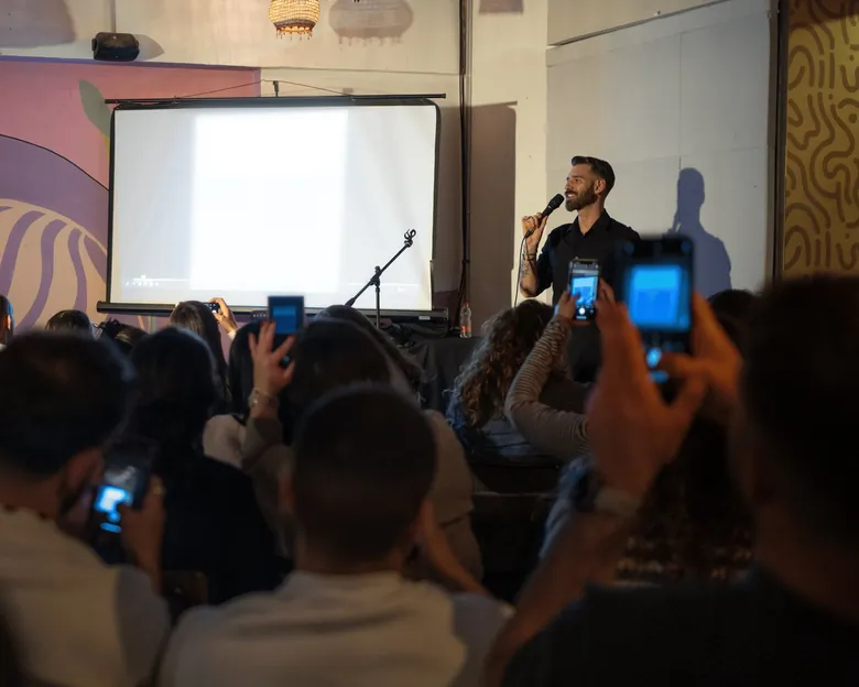 אוסקר מלכה מדבר מול אולם מלא, ובקדמת התמונה אנשים מהקהל מצלמים אותו בטלפונים